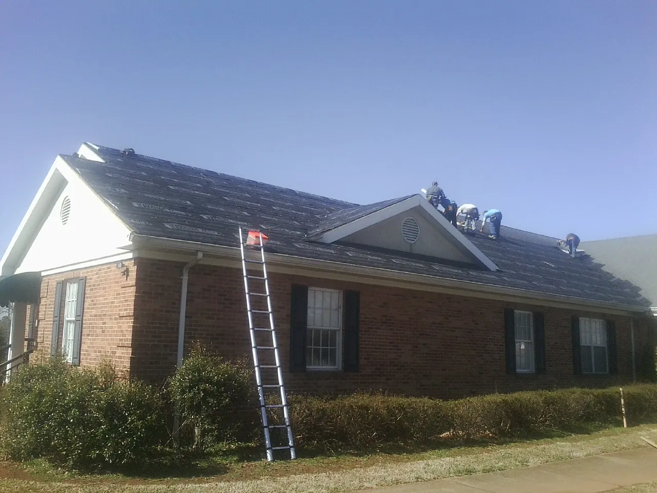 Crew tearing shingles off a brick ranch roof with a ladder set — Emergency Roof Repair work by our crew in Bloomington
